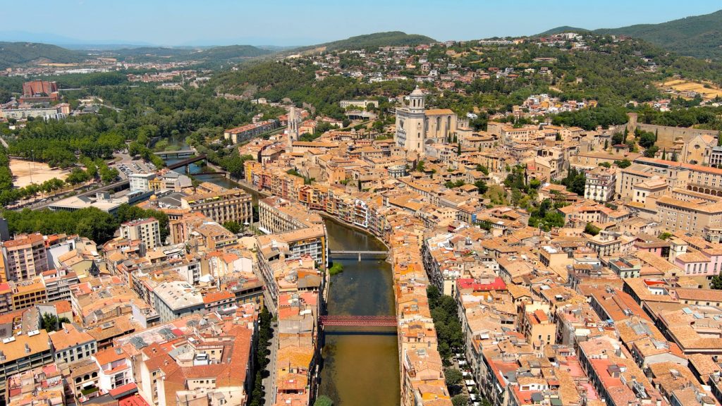 Eine weite Luftaufnahme zeigt den gewundenen Fluss Onyar, der sich durch das dichte Häusermeer von Girona bis zur Kathedrale schlängelt.