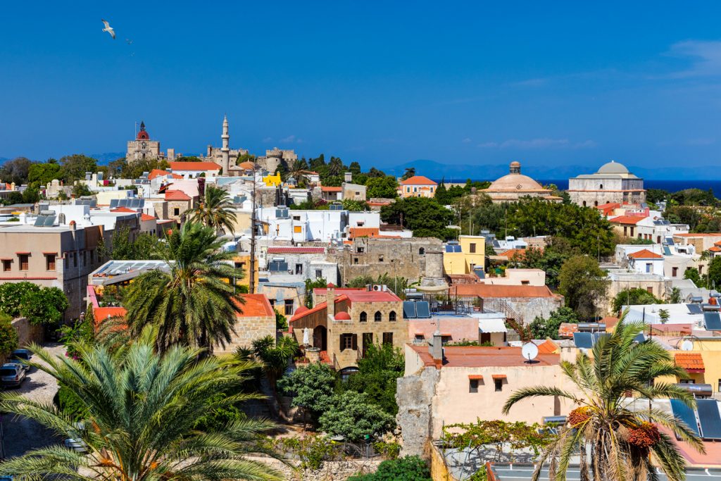 Eine weite Ansicht über zahlreiche helle Häuser mit flachen Dächern, durchsetzt von Palmen, Minaretten und Kuppelbauten vor dem blauen Meer am Horizont.