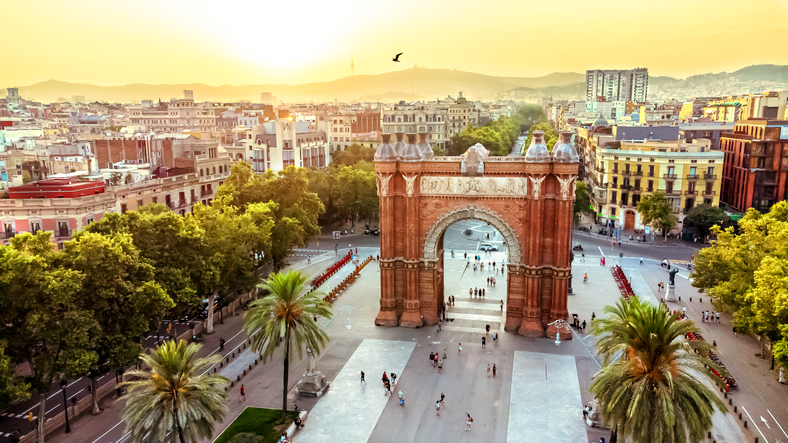 Eine Luftaufnahme des Arc de Triomf aus rotem Backstein am Ende einer von Palmen gesäumten Promenade.