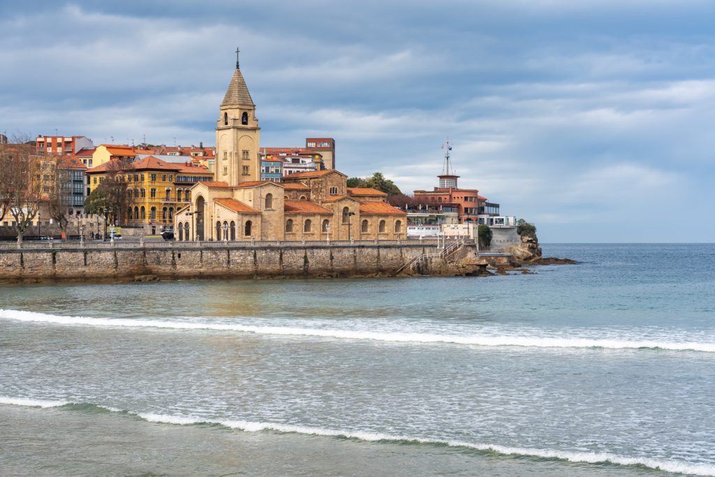 Eine markante Kirche mit Turm steht an der Uferpromenade vor sanften Wellen und der Stadtkulisse von Gijón unter wolkigem Himmel.