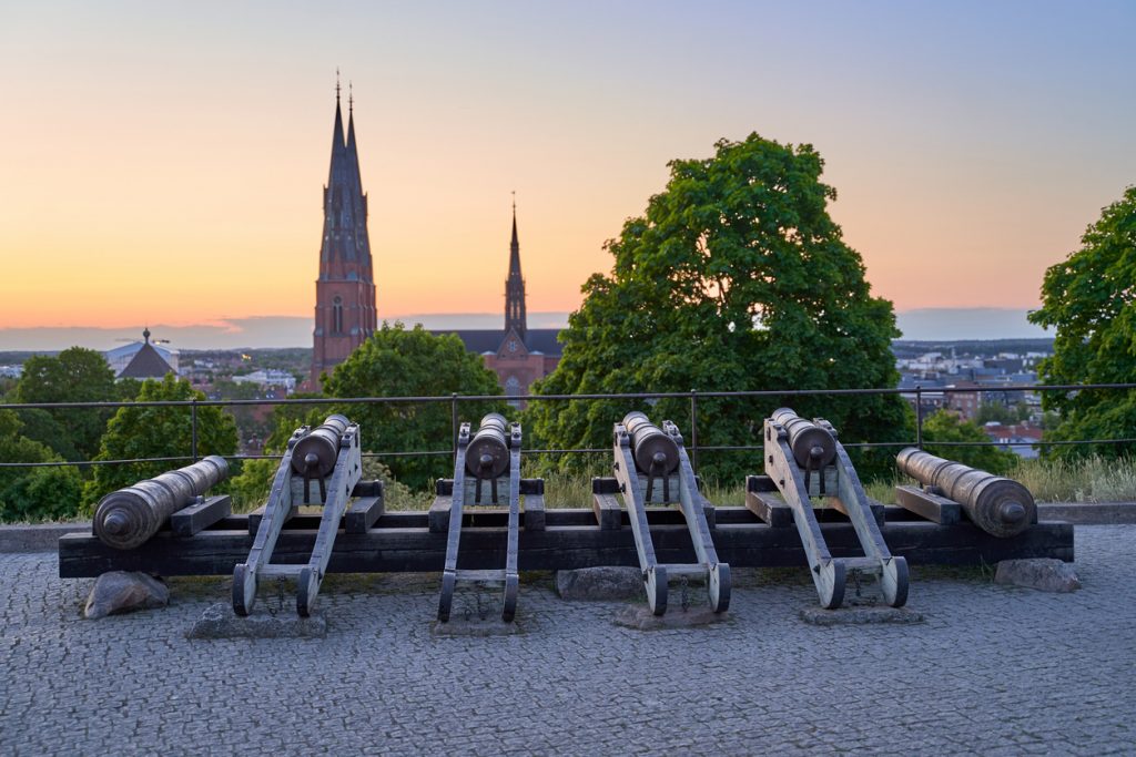 Historische Kanonen stehen auf einem Kopfsteinpflasterplatz vor der Silhouette des Doms von Uppsala bei Sonnenuntergang.