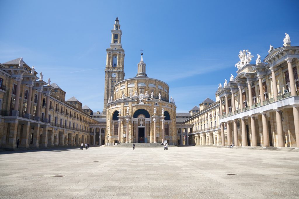 Ein weitläufiger steinerner Platz liegt vor einem imposanten historischen Bau mit Rundfassade, hohem Turm und langen Arkadengängen in Gijón.