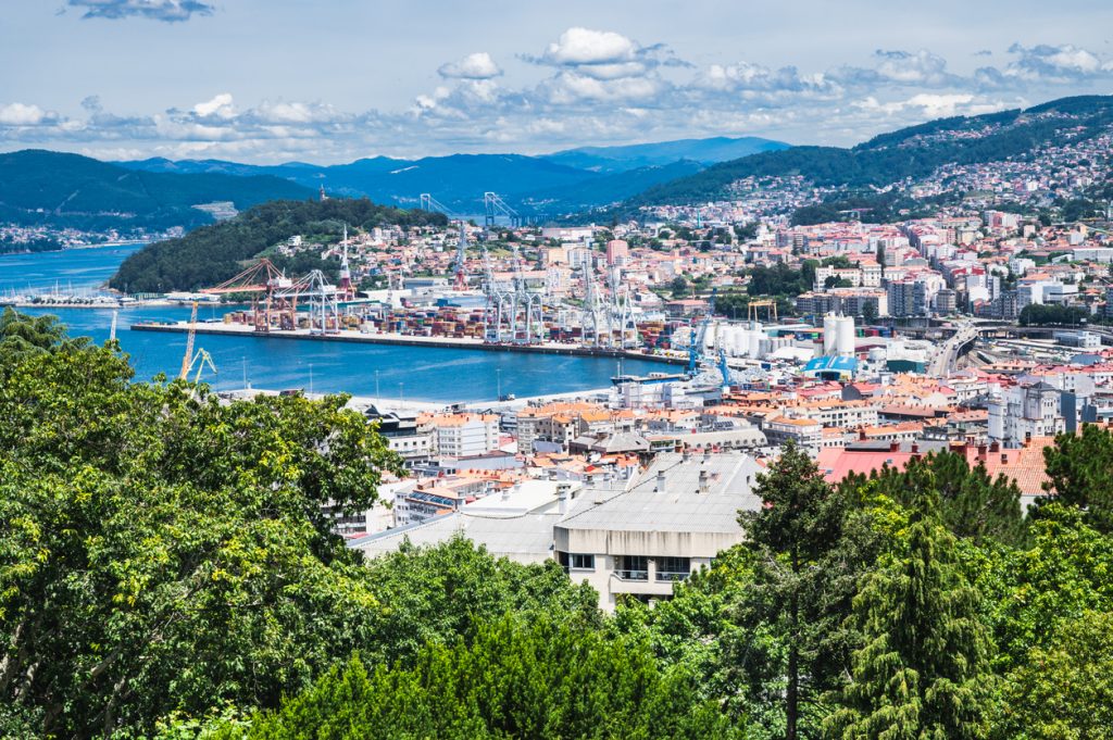 Ein weiter Ausblick von einem bewaldeten Hügel auf die Hafenanlagen mit Kränen, die Stadt und die dahinterliegende Bergkette unter blauem Himmel.