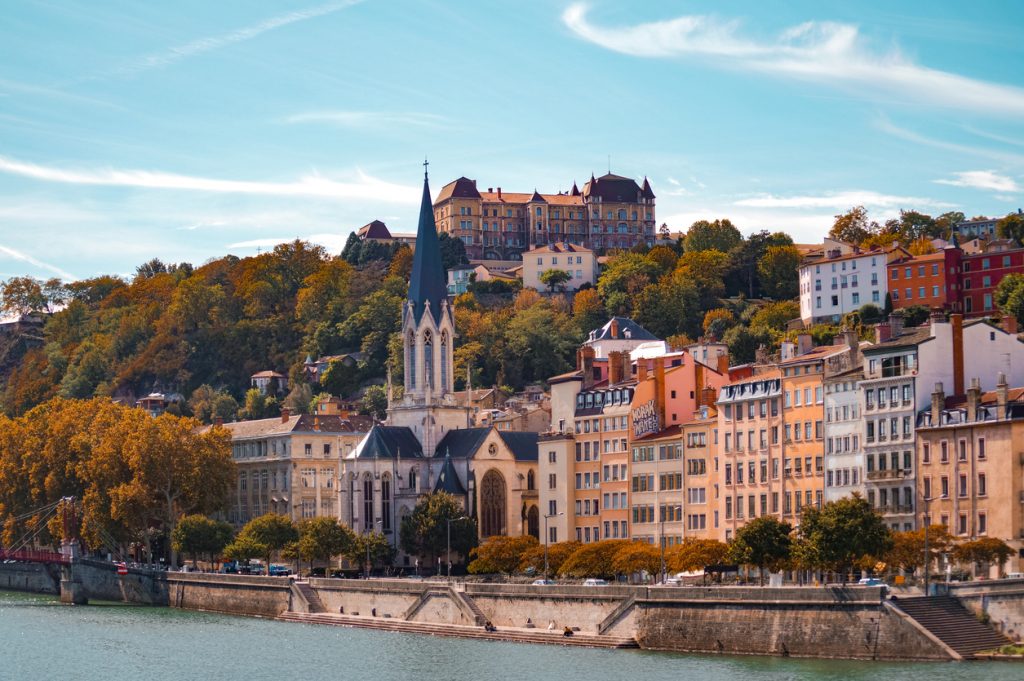 Blick auf Lyon mit bunten Fassaden am Fluss und einer Kirche auf dem Hügel.