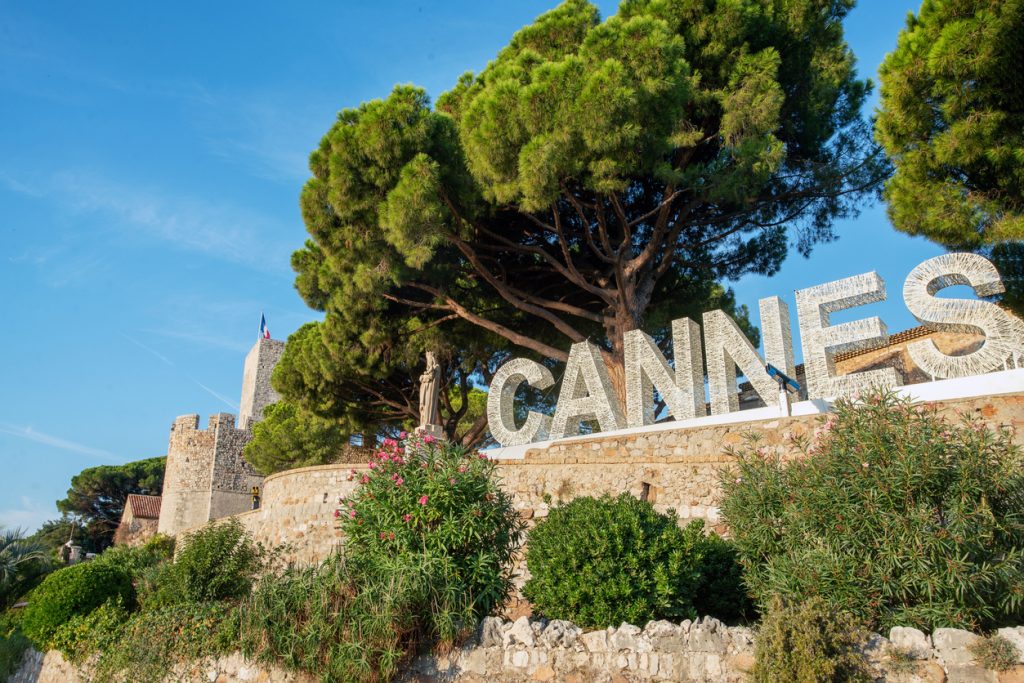 Große weiße Buchstaben bilden das Wort "CANNES" vor einer Kulisse aus Pinienbäumen und der alten Stadtmauer mit einer Marienstatue.