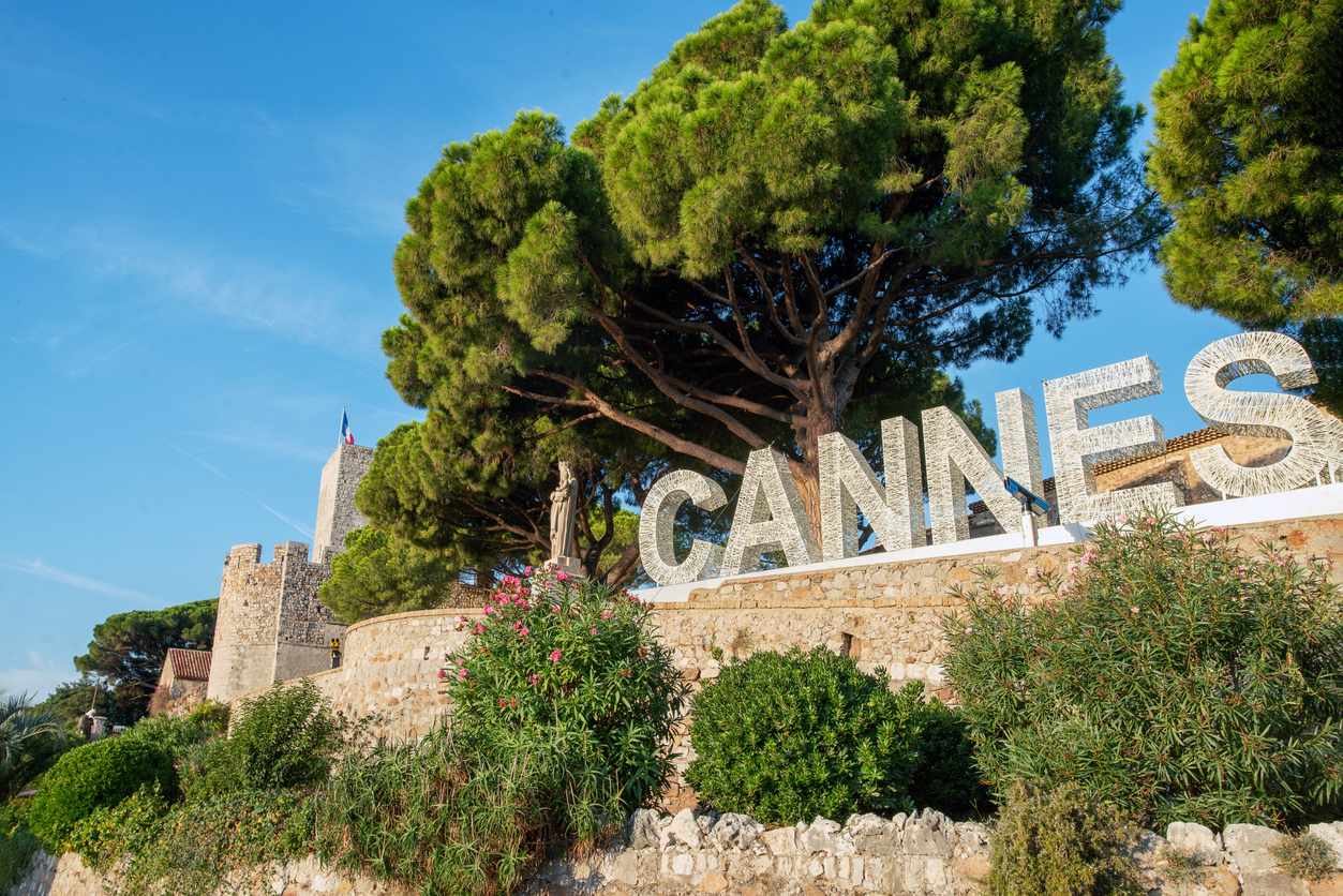 Große weiße Buchstaben bilden das Wort "CANNES" vor einer Kulisse aus Pinienbäumen und der alten Stadtmauer mit einer Marienstatue.