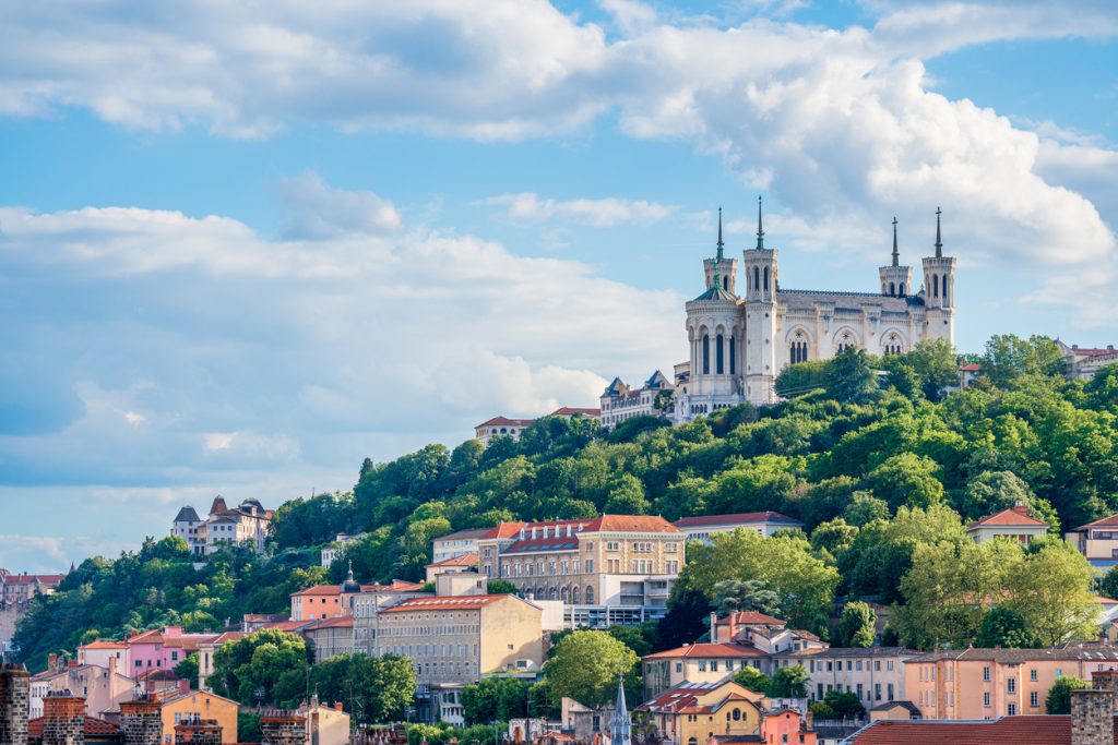 Blick auf die Basilika Notre-Dame de Fourvière auf dem Hügel über Lyon mit Wolken und grünen Hängen.
