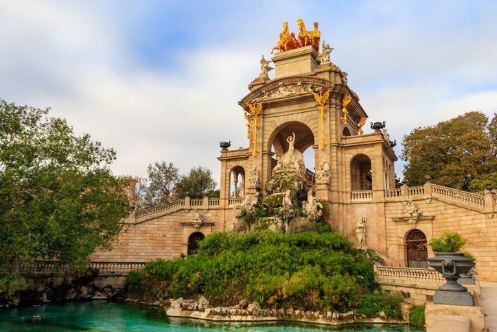 Ein monumentaler Springbrunnen mit goldenen Statuen und einer Quadriga auf der Spitze steht vor einem künstlichen See.