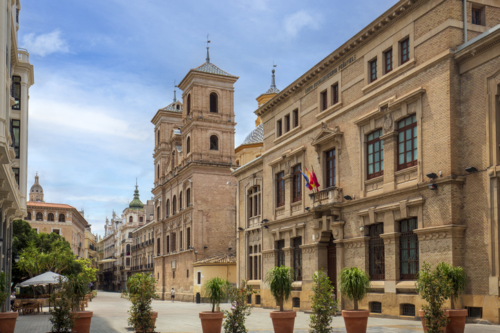Eine ruhige Straße im historischen Zentrum von Murcia, gesäumt von imposanten Backstein- und Sandsteingebäuden mit kunstvollen Verzierungen unter strahlend blauem Himmel.