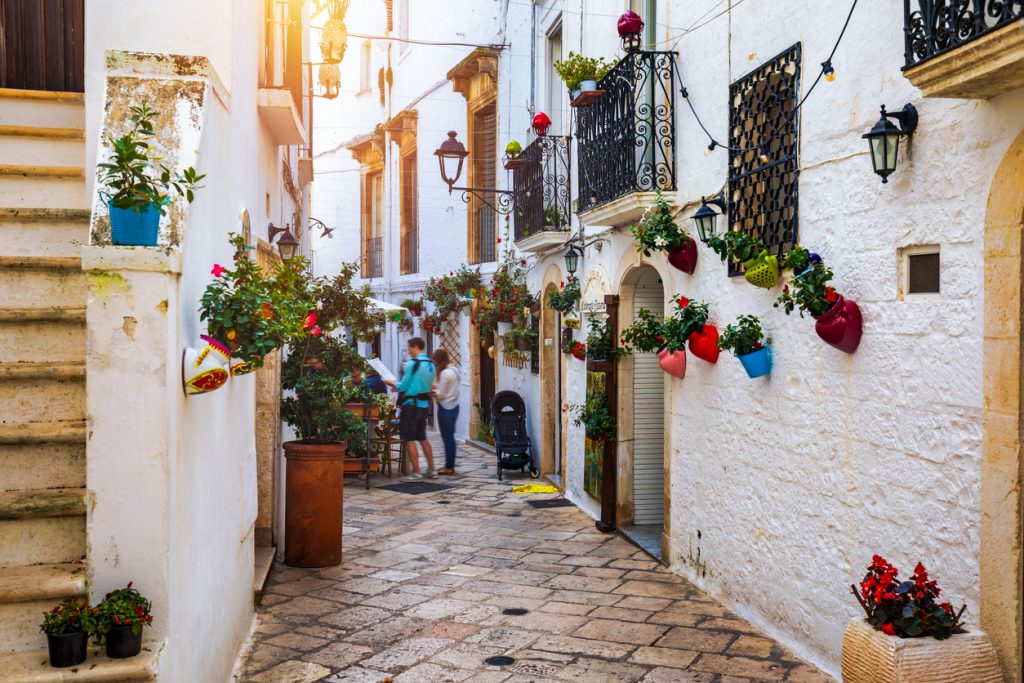 Eine weiß gekalkte Gasse mit Treppenstufen, an deren Wänden bunte Keramikgefäße in Herzform und andere Töpfe mit blühenden Pflanzen hängen.