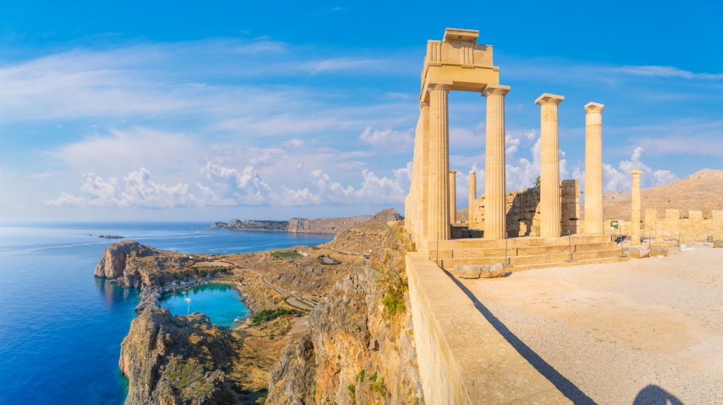 Mehrere gut erhaltene dorische Säulen stehen am Rand eines Abgrunds vor einem strahlend blauen Himmel; tief unten ist eine herzförmige Meeresbucht mit türkisblauem Wasser erkennbar.