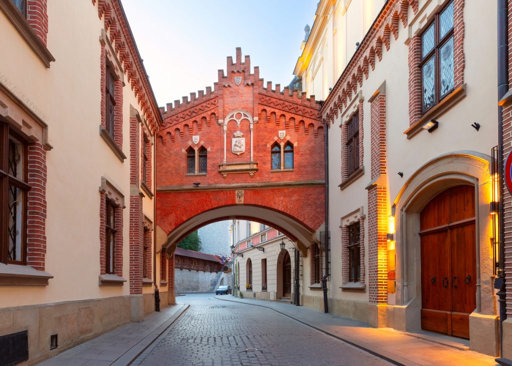 Ein markanter Torbogen aus rotem Backstein in Krakau verbindet zwei historische Gebäude über einer gepflasterten Gasse.