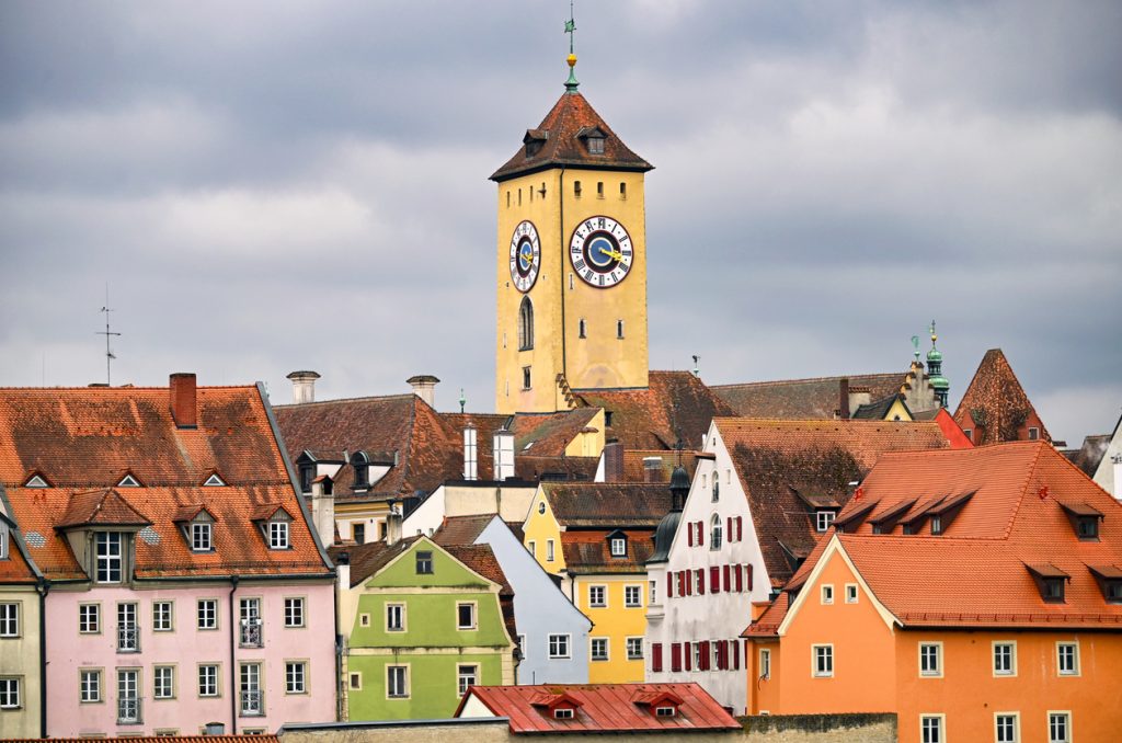 Blick über die bunten Fassaden und roten Ziegeldächer von Regensburg zum markanten Rathausturm mit seiner großen Uhr.