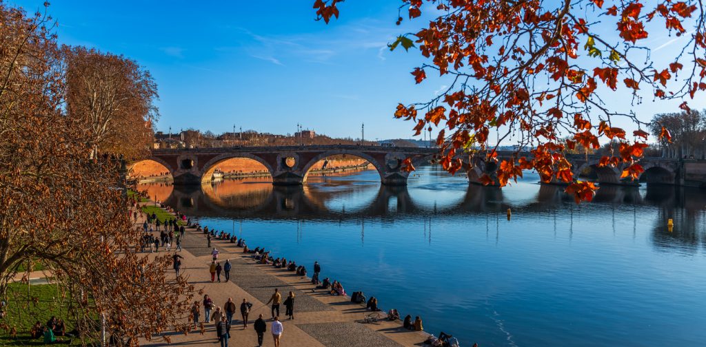 Menschen spazieren am sonnigen Flussufer entlang, im Hintergrund überspannt die Pont Neuf das Wasser.