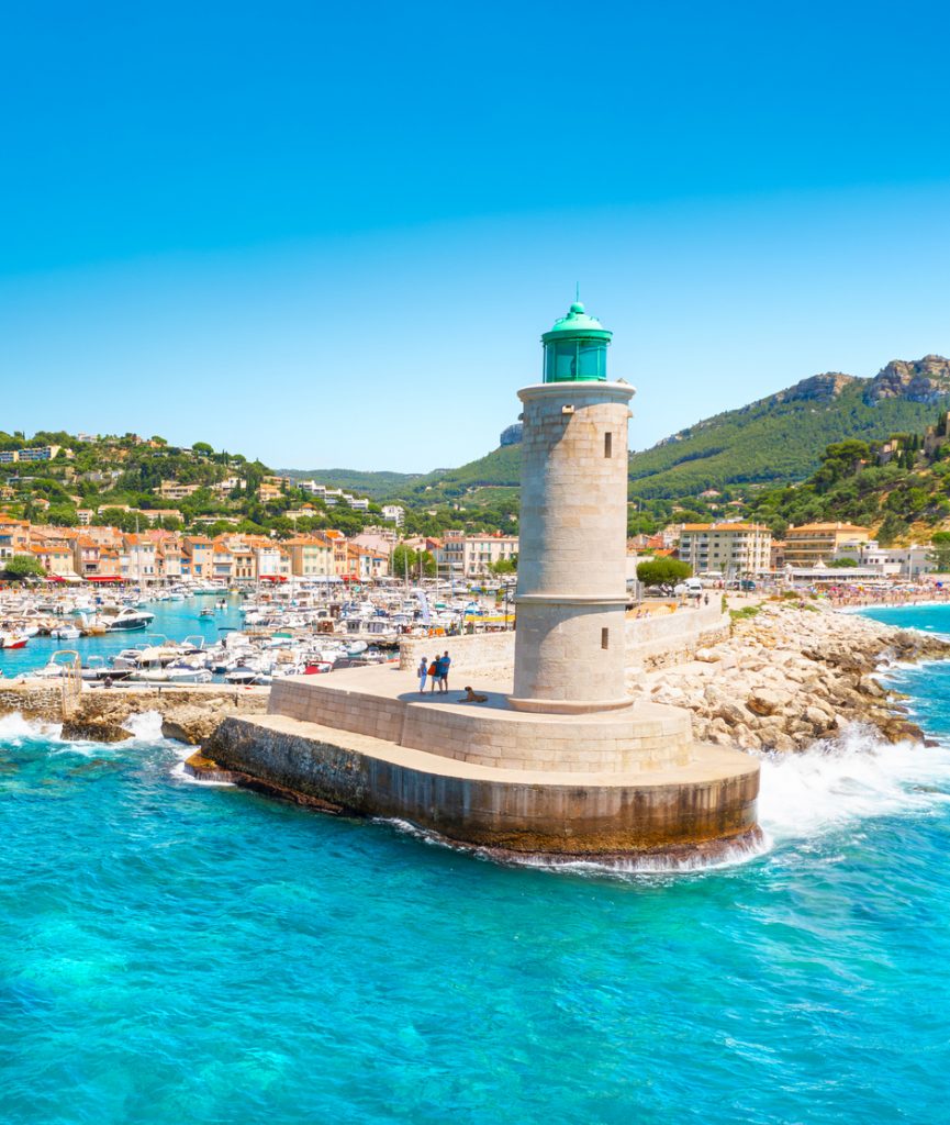 Ein heller Steinturm mit grüner Spitze steht auf einer Hafenmauer vor einer Kulisse aus Bergen, türkisfarbenem Wasser und vielen Segelbooten.