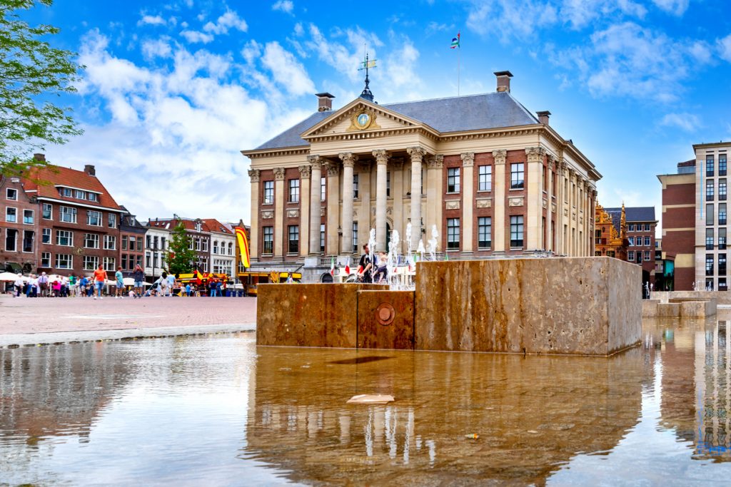 Ein klassizistisches Gebäude mit Säulen steht an einem belebten Platz hinter einem modernen Brunnen.