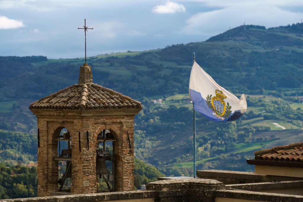 Die blau-weiße Nationalflagge weht vor einem historischen Glockenturm und bewaldeten Bergen
