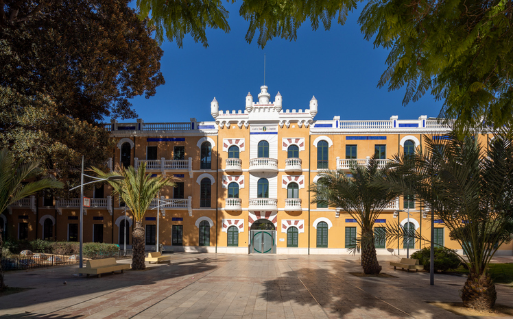 Ein prächtiges, gelbes Gebäude mit hufeisenförmigen Fenstern und weißen Ornamenten steht an einem sonnigen Platz mit Palmen.