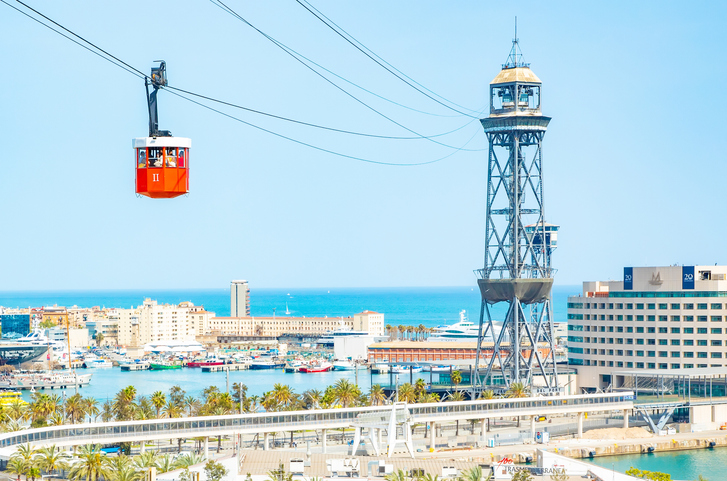 Eine rote Kabine der Hafenseilbahn schwebt vor dem stählernen Torre Jaume I und dem tiefblauen Meer.