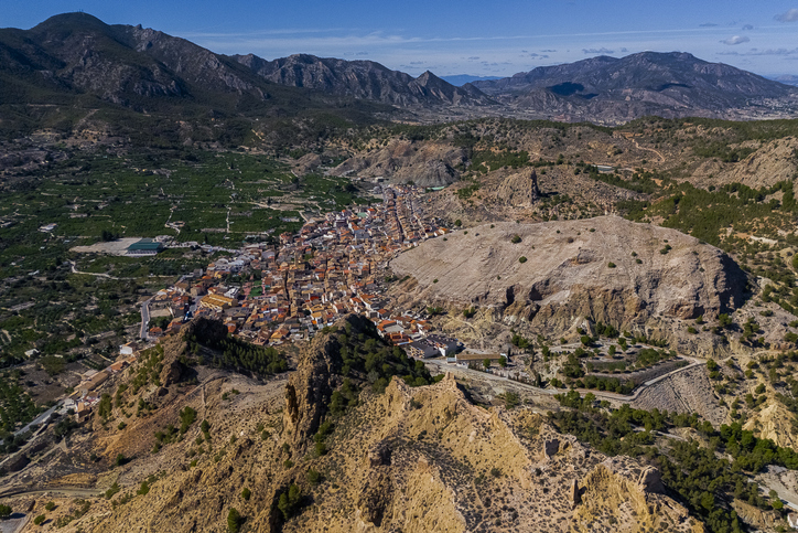 Eine weite Luftaufnahme zeigt eine Stadt, die sich an karge, zerklüftete Berge schmiegt, umgeben von fruchtbaren grünen Plantagen.
