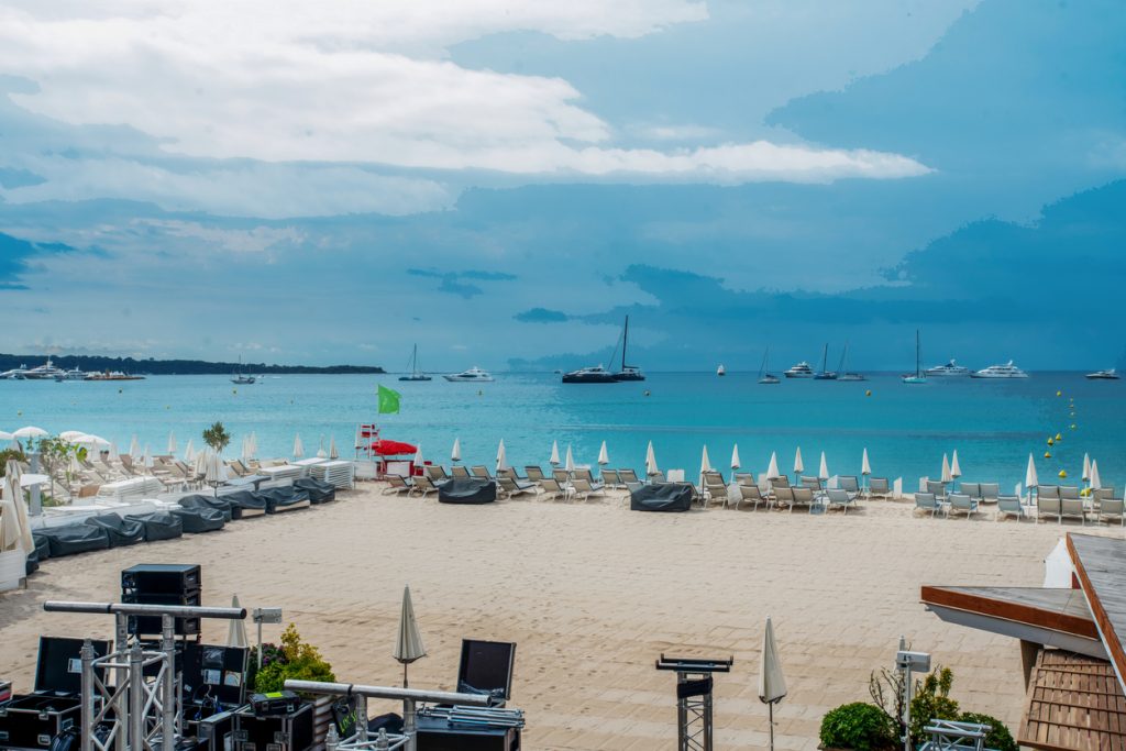 Ein heller Sandstrand mit ordentlich aufgereihten Liegestühlen und weißen Sonnenschirmen unter einem bewölkten blauen Himmel; im Hintergrund ankern Yachten auf dem türkisfarbenen Wasser.