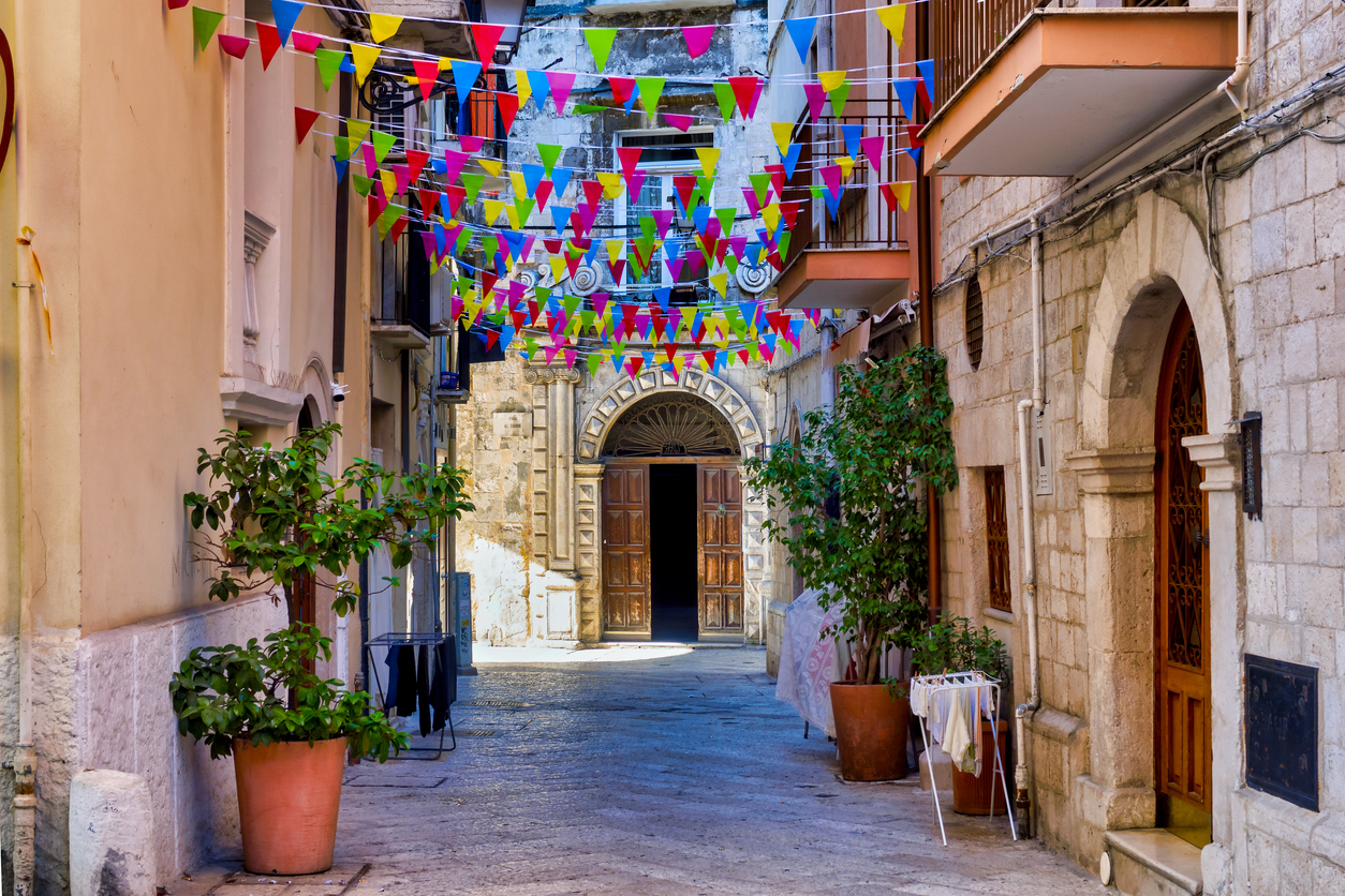 Eine schmale Gasse mit hellen Steinfassaden, über der zahlreiche bunte Wimpelketten hängen; im Hintergrund ist ein verzierter Torbogen sichtbar.