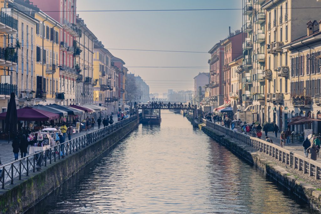 Blick entlang eines Kanals in Mailand mit Häuserfassaden, Menschen und Brücke in der Ferne.