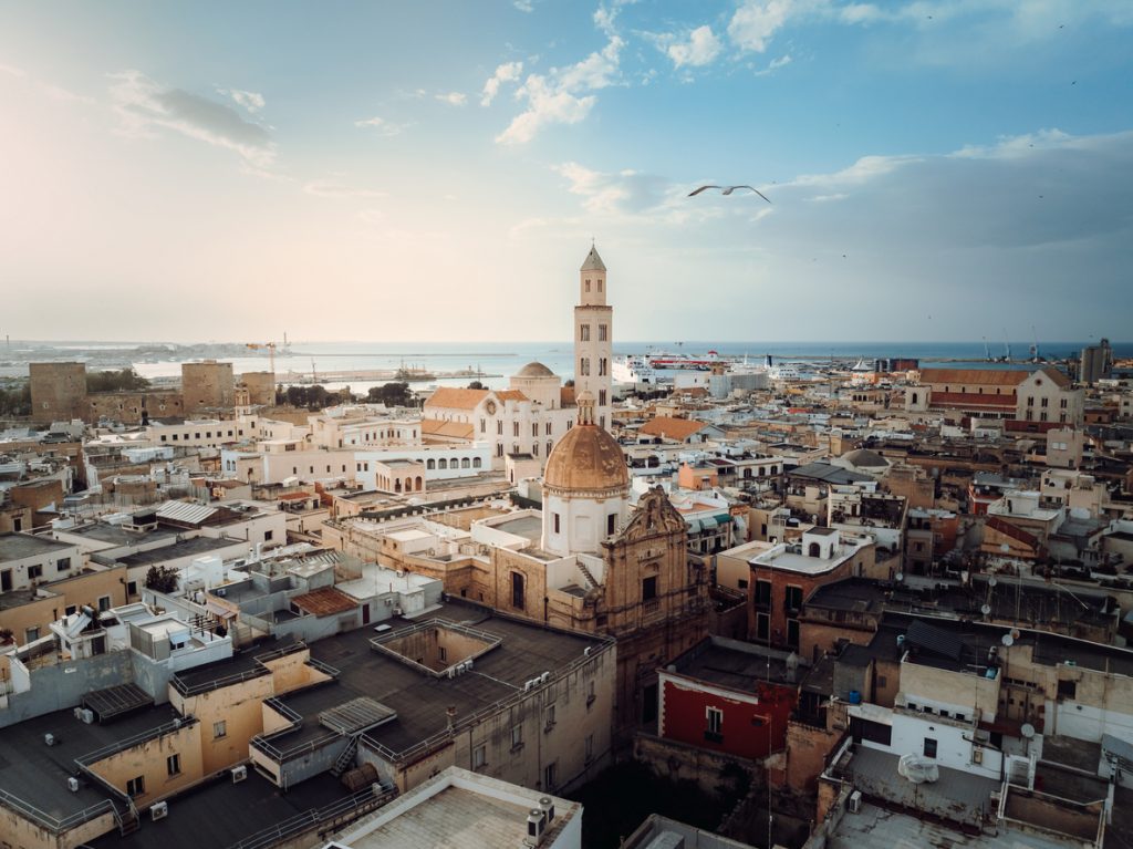 Eine Luftaufnahme der dichten Dachlandschaft der Stadt mit dem markanten Glockenturm und der Kuppel einer Kathedrale vor dem blauen Meer.