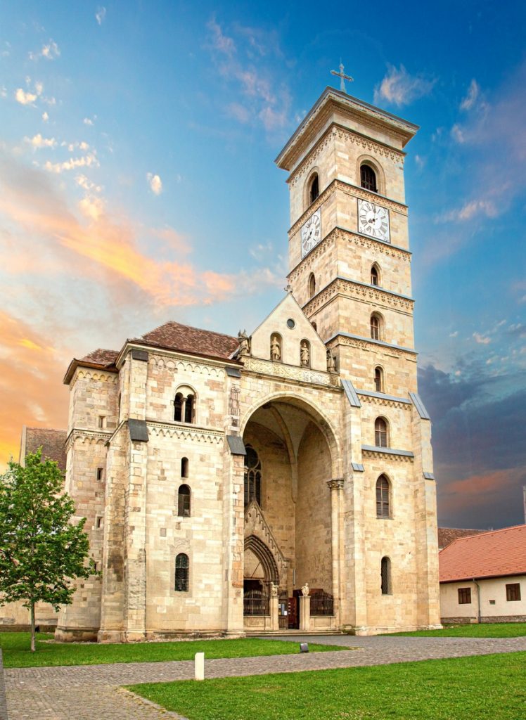 Die hohe Steinfassade einer romanischen Kathedrale mit einem markanten Uhrturm vor einem farbenfrohen Abendhimmel.