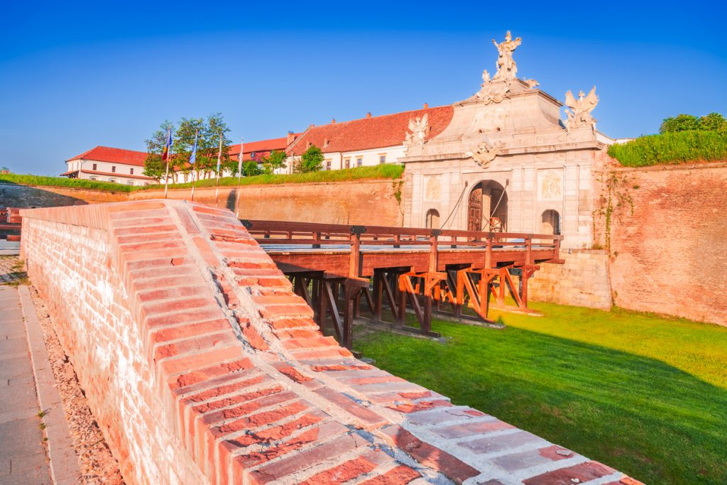 Ein monumentales Steintor mit barocken Skulpturen und einer hölzernen Zugbrücke bei strahlendem Sonnenschein.