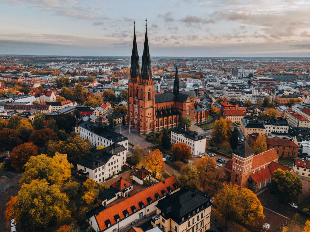 Eine Drohnenaufnahme zeigt den roten Backsteinbau des Doms von Uppsala inmitten herbstlich bunter Bäume.