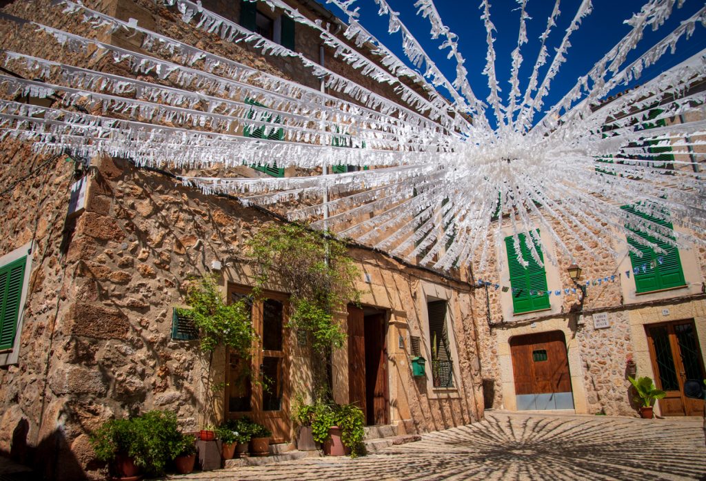 Zahlreiche weiße Papierschnüre sind strahlenförmig über eine schmale Gasse mit traditionellen Natursteinhäusern und grünen Fensterläden gespannt.