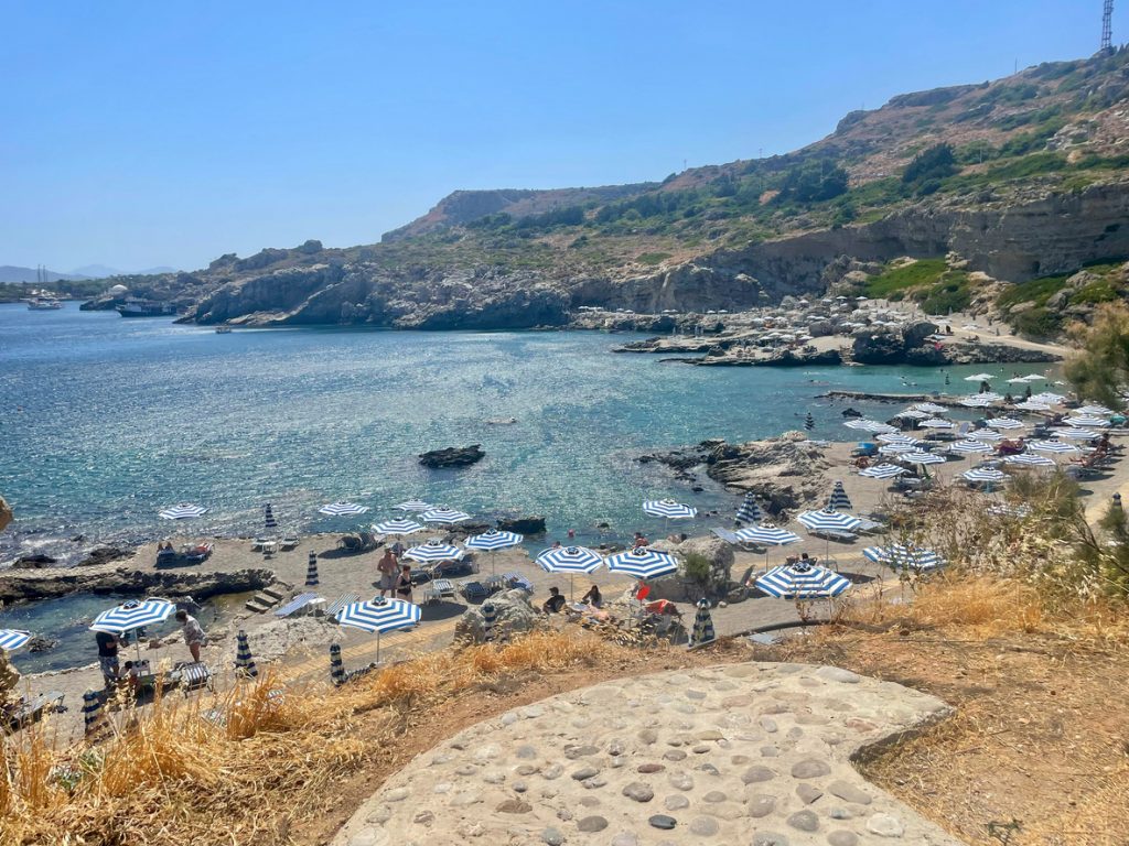 Ein Blick von einer Anhöhe auf eine felsige Meeresbucht mit zahlreichen blau-weiß gestreiften Sonnenschirmen und Liegen auf den flachen Felsen am Ufer.