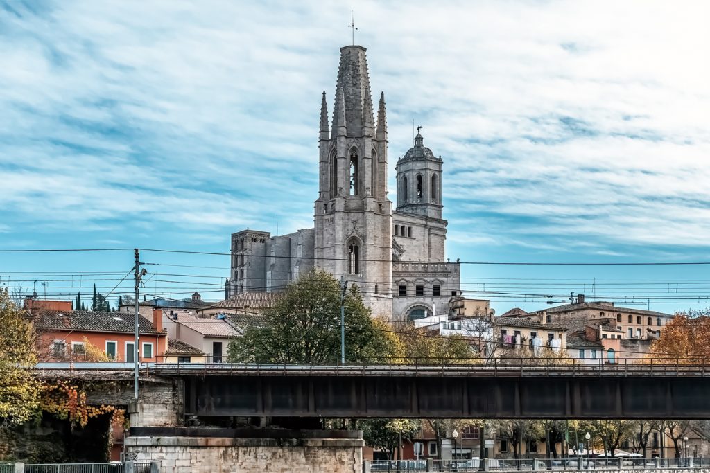 Der schlanke Turm der Basilika Sant Feliu in Girona ragt hinter einer Eisenbahnbrücke in den bewölkten Himmel.