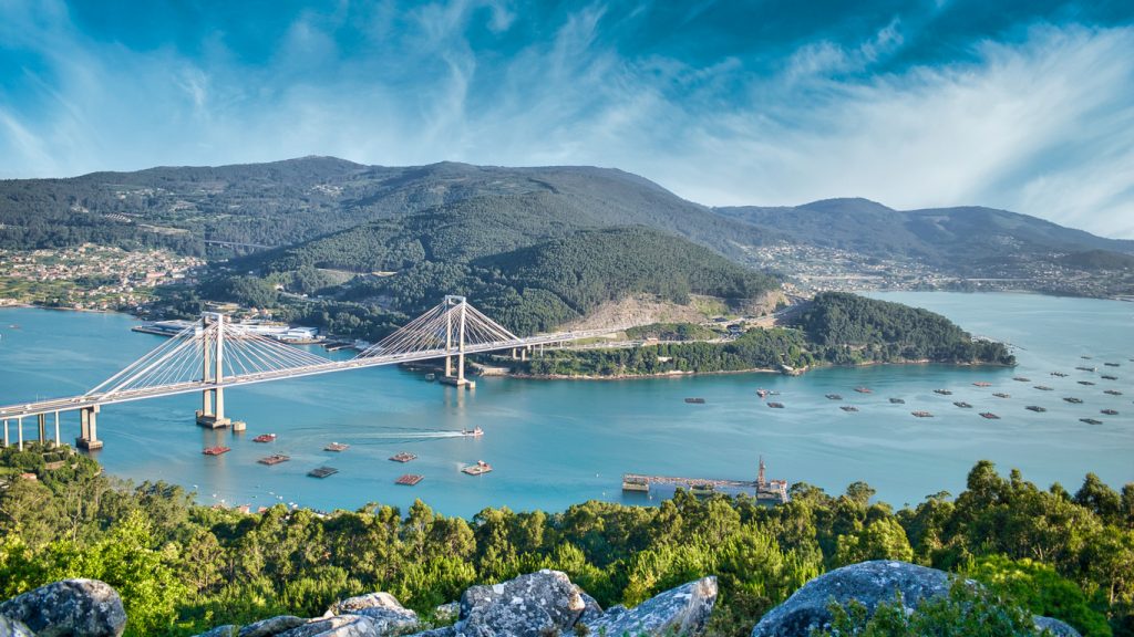 Eine große Schrägseilbrücke spannt sich über eine weite Meeresbucht und verbindet zwei bergige Küstenabschnitte unter einem dynamischen Wolkenhimmel.