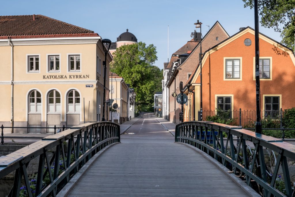 Eine Holzbrücke führt über einen Kanal auf eine Straße mit farbenfrohen Häusern und dem Schloss Uppsala im Hintergrund.