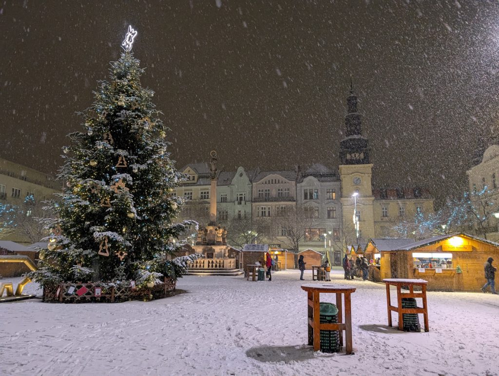 Verschneiter Weihnachtsmarkt bei Nacht mit großem Baum, Buden und fallenden Schneeflocken.