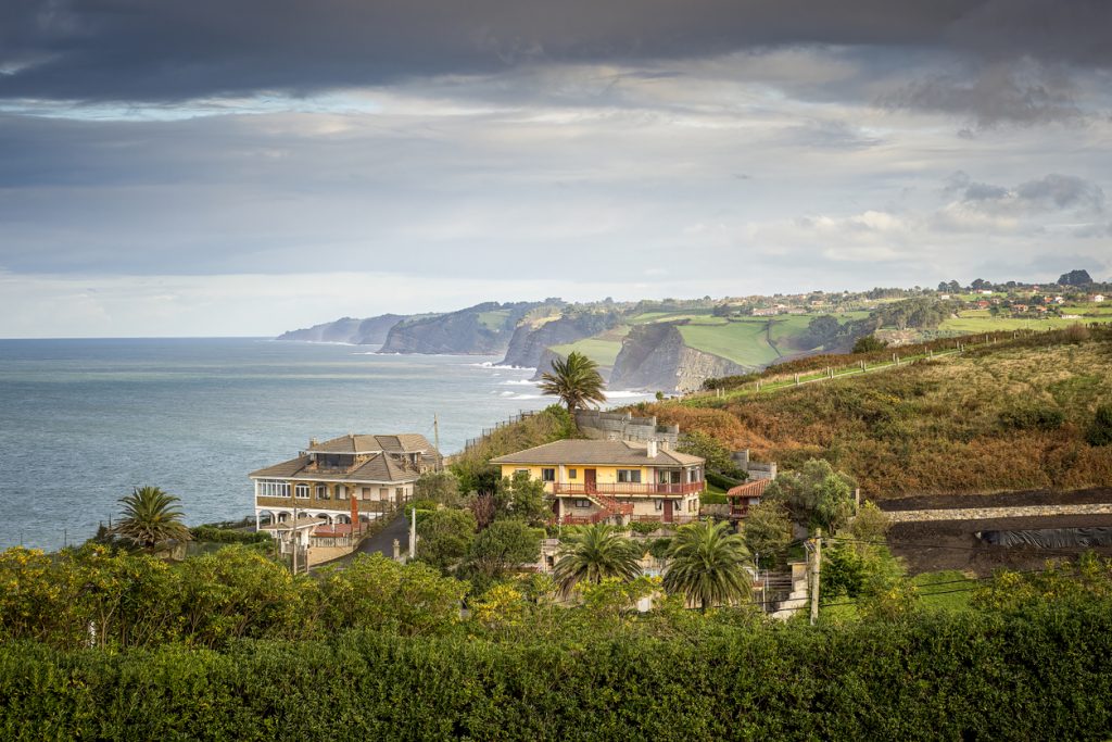 Häuser, Palmen, grüne Felder und steile Klippen ziehen sich entlang der Küste, während das Meer unter dramatischem Himmel vor Gijón liegt.