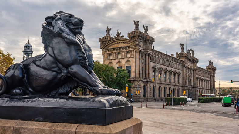 Eine große Löwenstatue aus dunklem Metall wacht vor dem prächtigen historischen Gebäude der Hafenbehörde in Barcelona.