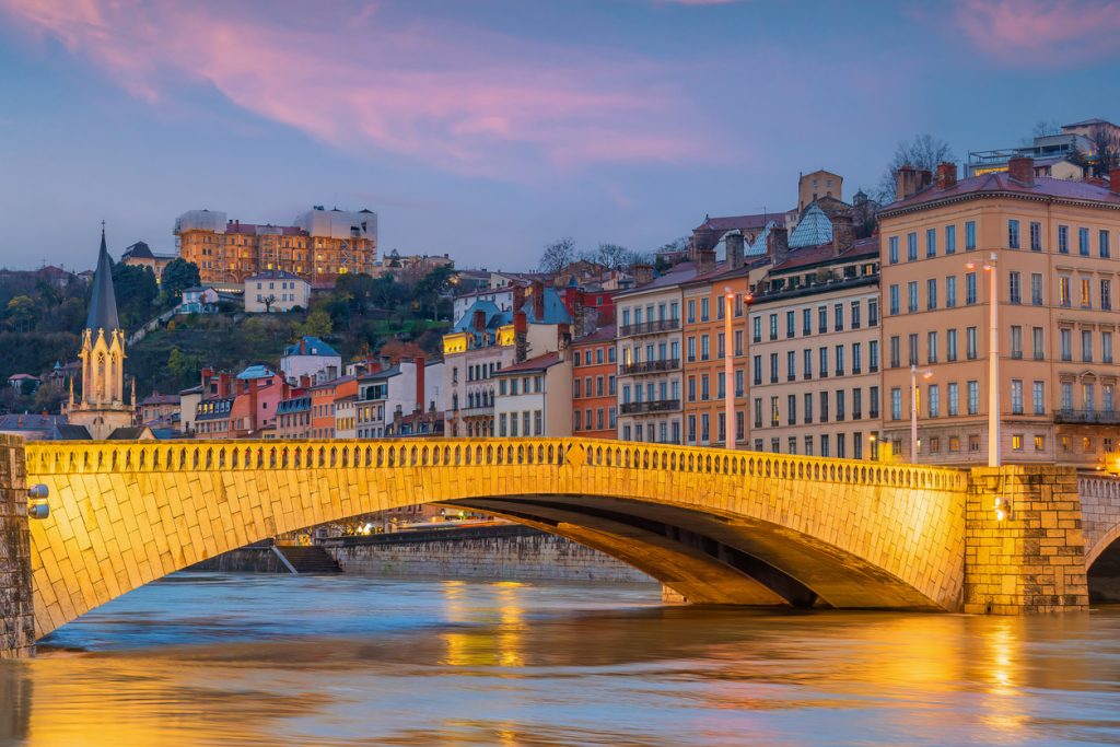 Beleuchtete Steinbrücke über den ruhigen Fluss, dahinter bunte Häuserzeilen und ein violettblauer Himmel in der Dämmerung.