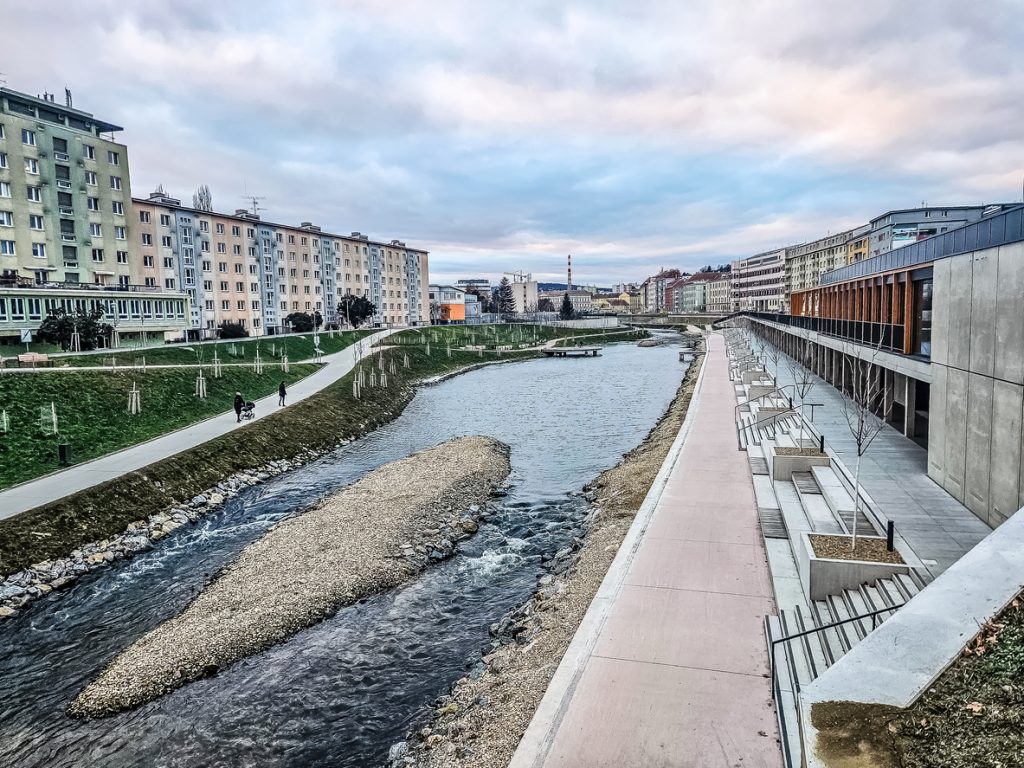 Flusslauf mit Spazierwegen, Treppenanlagen und Wohnblöcken unter bewölktem Himmel.