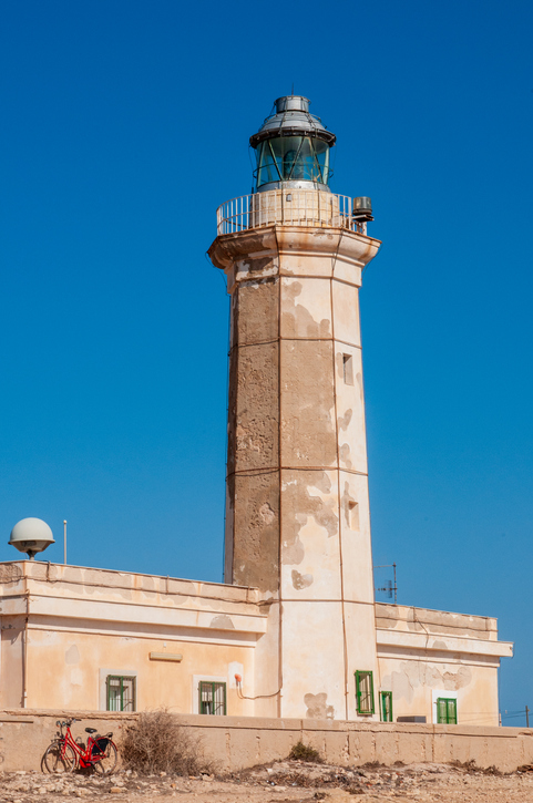 Ein hoher, sandfarbener Leuchtturm steht vor einem tiefblauen, wolkenlosen Himmel, davor parkt ein rotes Fahrrad.