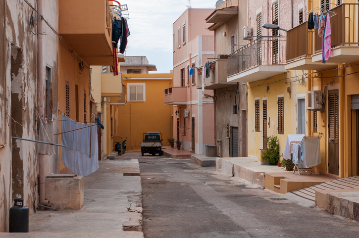 Eine schmale Gasse mit ockerfarbenen Häusern, Wäscheleinen auf den Balkonen und einem kleinen Dreirad-Transporter.