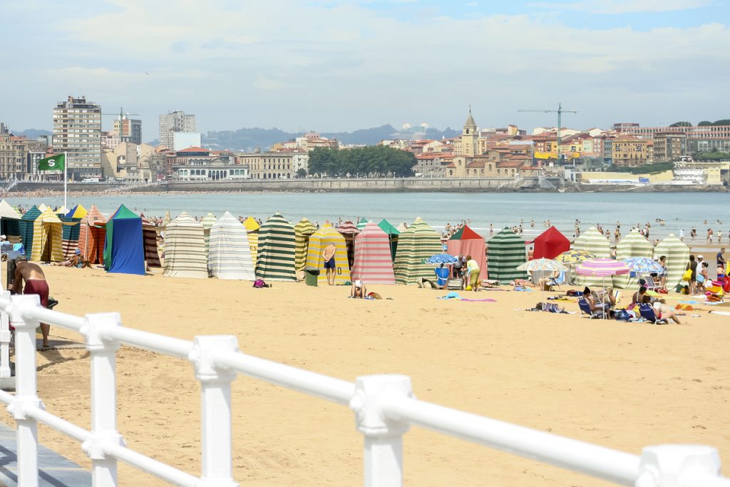 Ein breiter Sandstrand mit vielen bunten Strandkabinen, Badenden und Blick auf die Küste und Häuser von Gijón erstreckt sich unter hellem Himmel.