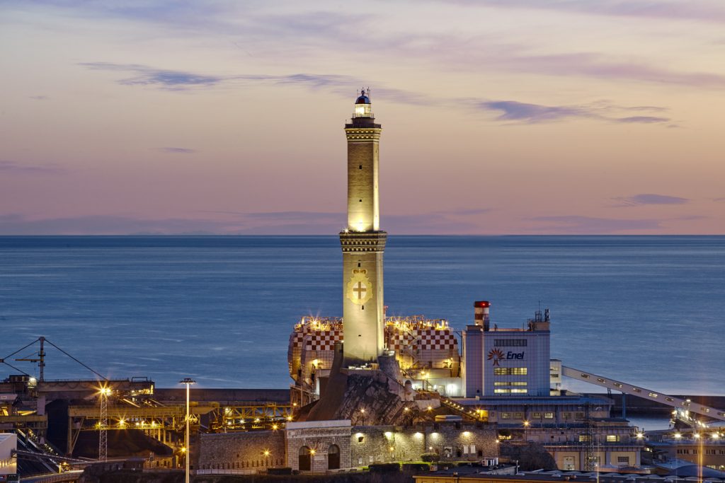 Der beleuchtete Leuchtturm von Genua steht majestätisch am Hafenrand vor einem violett-goldenen Abendhimmel.