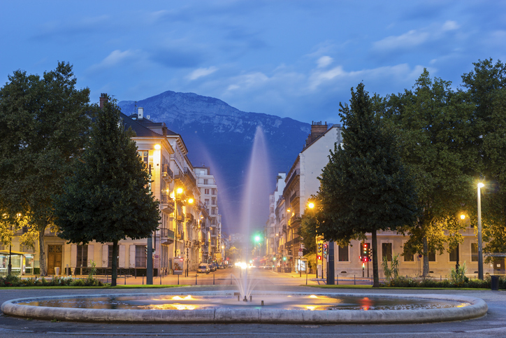 Ein beleuchteter Springbrunnen im Vordergrund einer von Bäumen gesäumten Stadtstraße bei Dämmerung, im Hintergrund ragt ein massiver Berg auf.