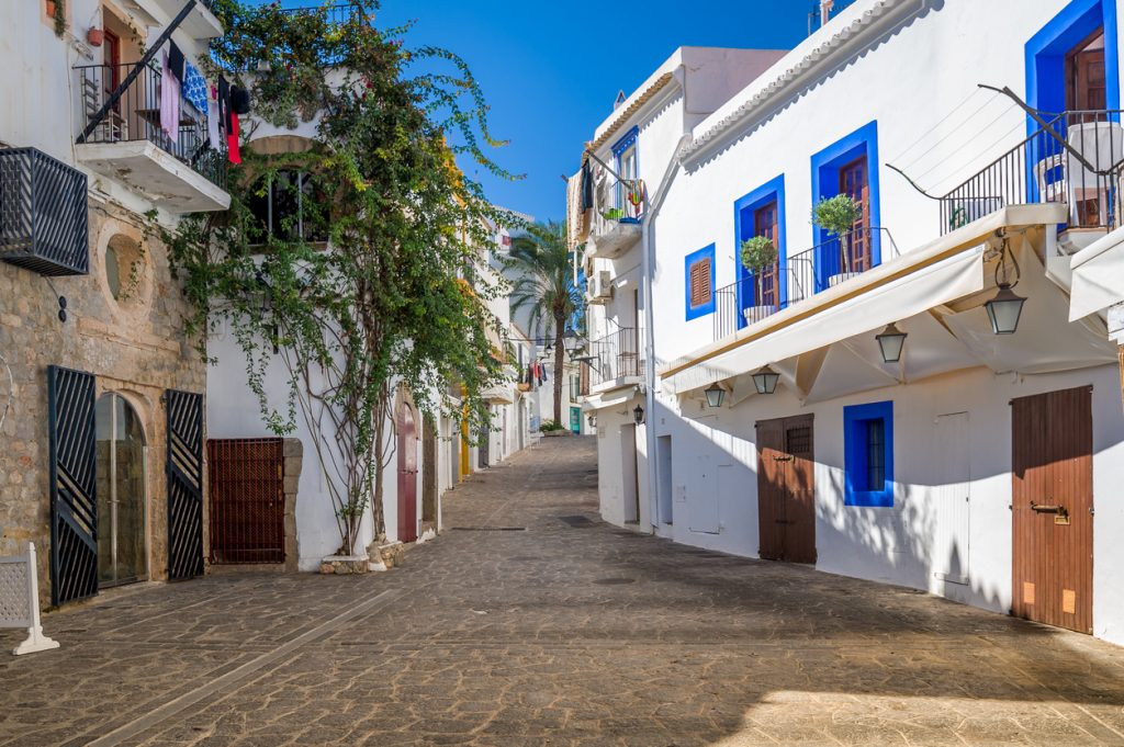 Schmale Straße auf Ibiza mit weißen Häusern, blauen Fensterrahmen, Balkonen und Sonnenschatten.