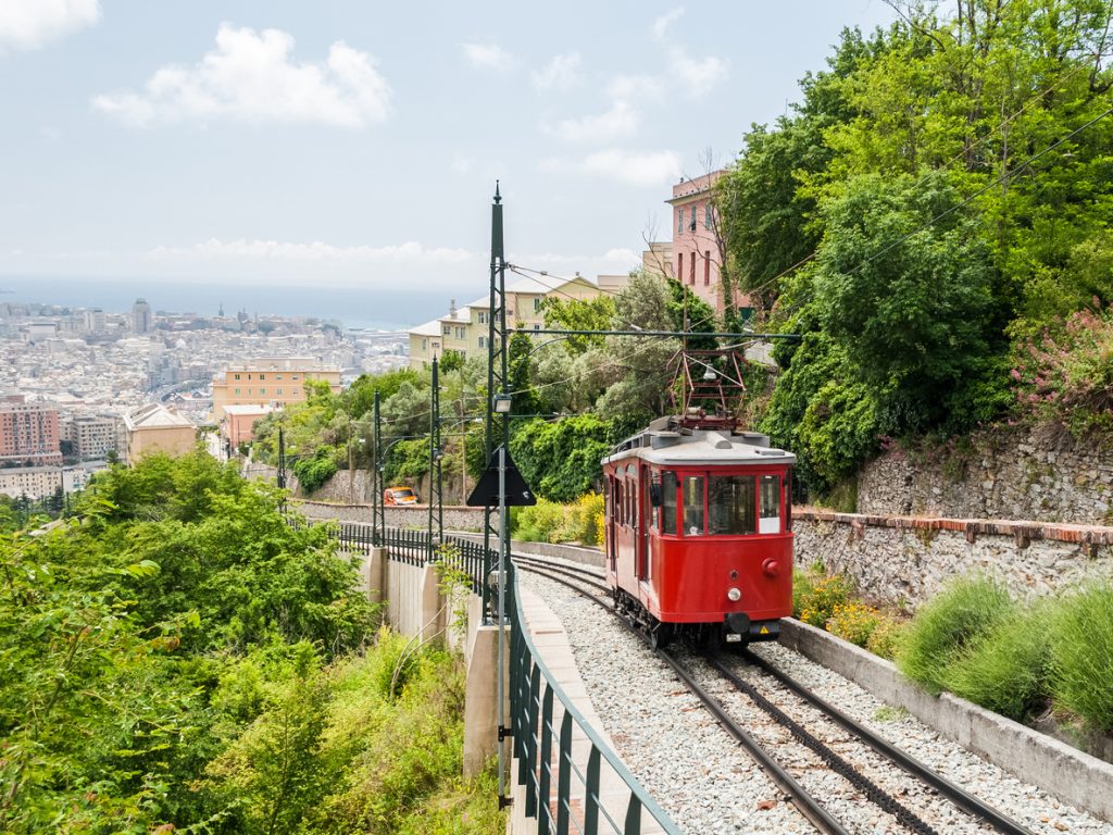 Eine historische rote Bahn fährt auf Schienen einen grünen Hügel in Genua hinauf, mit der Stadt und dem Meer im Hintergrund.