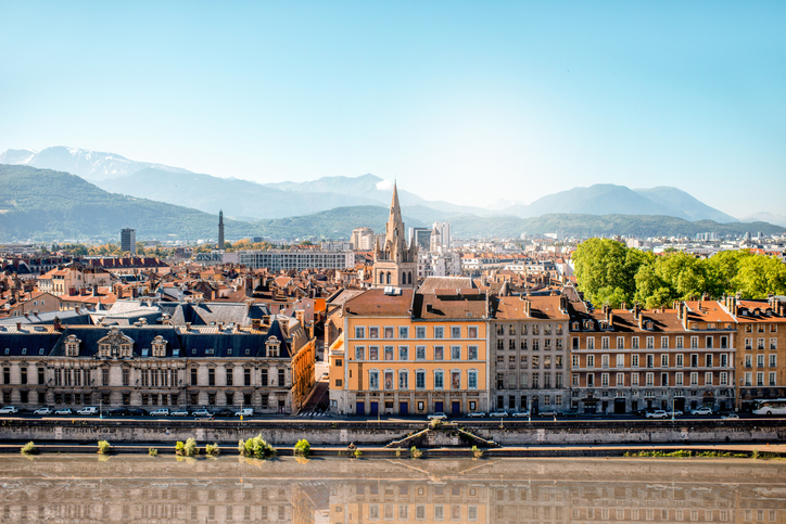 Blick von oben auf die dichte Bebauung von Grenoble mit einer zentralen Kirche und dem Fluss Isère vor einer weiten Gebirgskette.