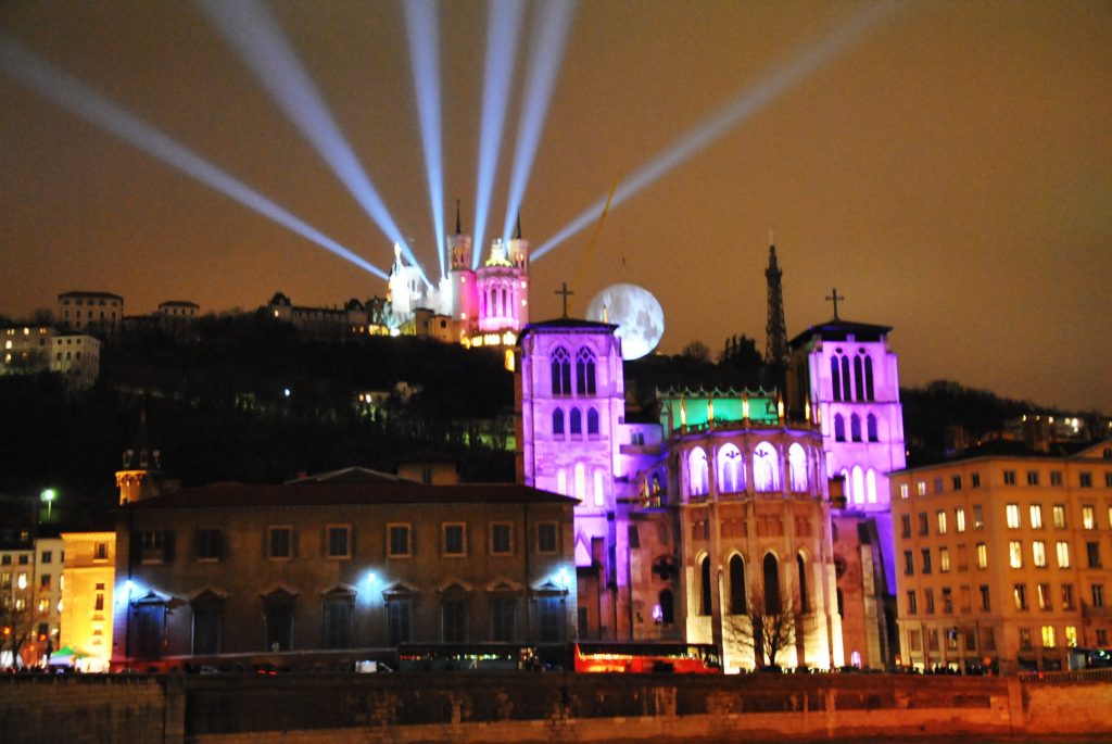 Angestrahlte Kirche in kräftigen Farben mit Lichtkegeln in den Himmel und großem Mond im Hintergrund bei Nacht.